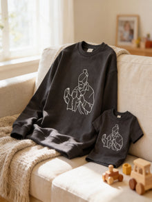 a gray sweatshirt with a white graphic design depicting a woman, and a smaller gray t-shirt with a similar white graphic design. The sweatshirt and t-shirt are placed on a couch, and there are some wooden toys in the foreground.