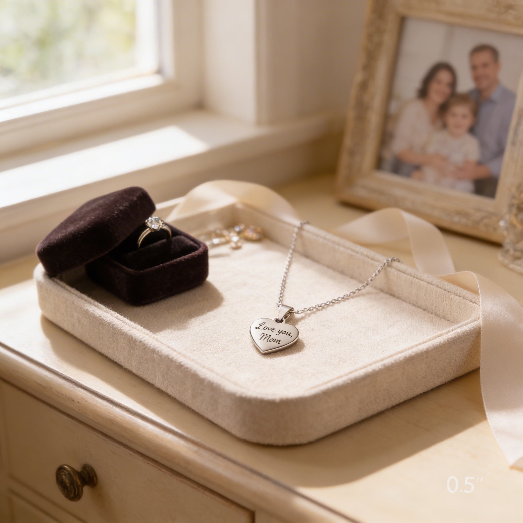A jewelry box containing a ring and a heart-shaped charm with the inscription "Love you Mom" is placed on a wooden dresser next to a framed photo of a family.