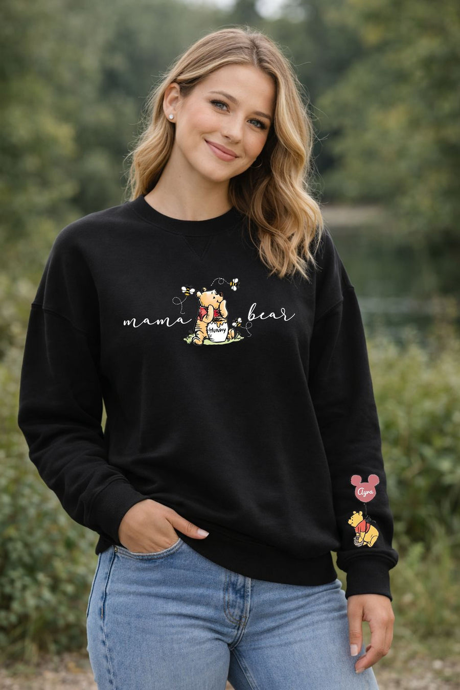 A smiling woman wearing a black sweatshirt with a Winnie the Pooh design and text printed on it, standing in a natural outdoor setting.