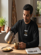 A man in a black sweatshirt is pouring a hot beverage from a silver teapot into a mug, with a plate of cookies and a newspaper on the table in front of him.