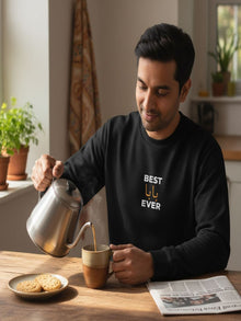 A man in a black sweatshirt is pouring a hot beverage from a silver teapot into a mug, with a plate of cookies and a newspaper on the table in front of him.