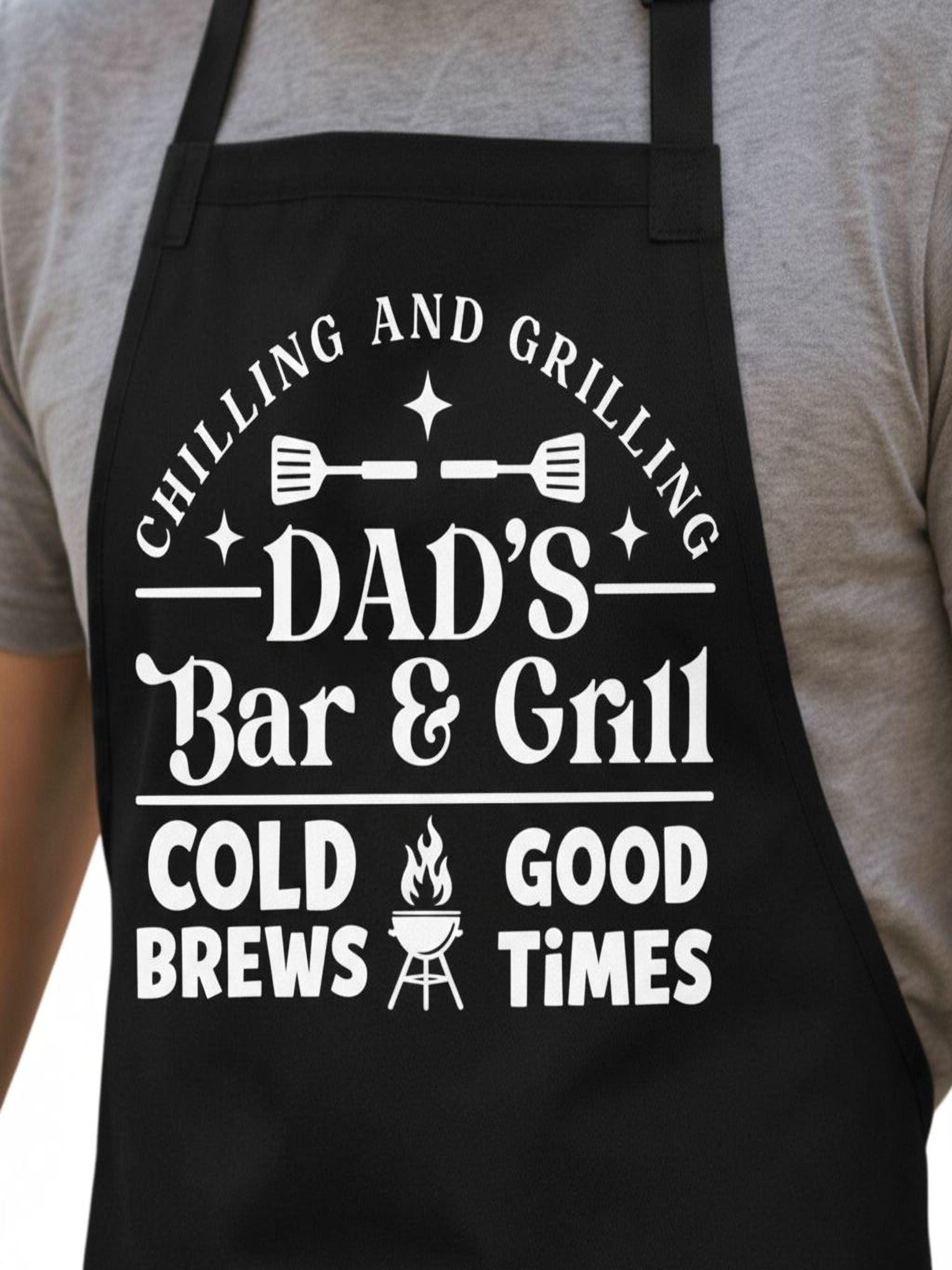 Dad’s Bar and Grill apron featuring funny BBQ typography design worn by man grilling outdoors in backyard.