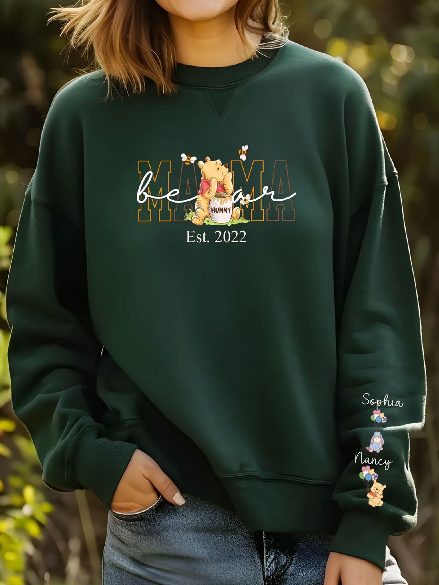 A woman wearing a green sweatshirt with a Winnie the Pooh design and text printed on it, standing in a natural outdoor setting with trees and foliage in the background.