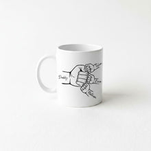 A white ceramic mug with a black and white illustration of two hands, one larger and one smaller, connected by a fist. The text "Daddy, Tyler, Liza, Nathan" is written above the illustration.