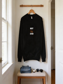 A black sweatshirt with the text "BEST U EVER" printed on it is hanging on a wooden hanger on a wall. Below the sweatshirt, there is a folded pair of jeans and a pair of sunglasses.