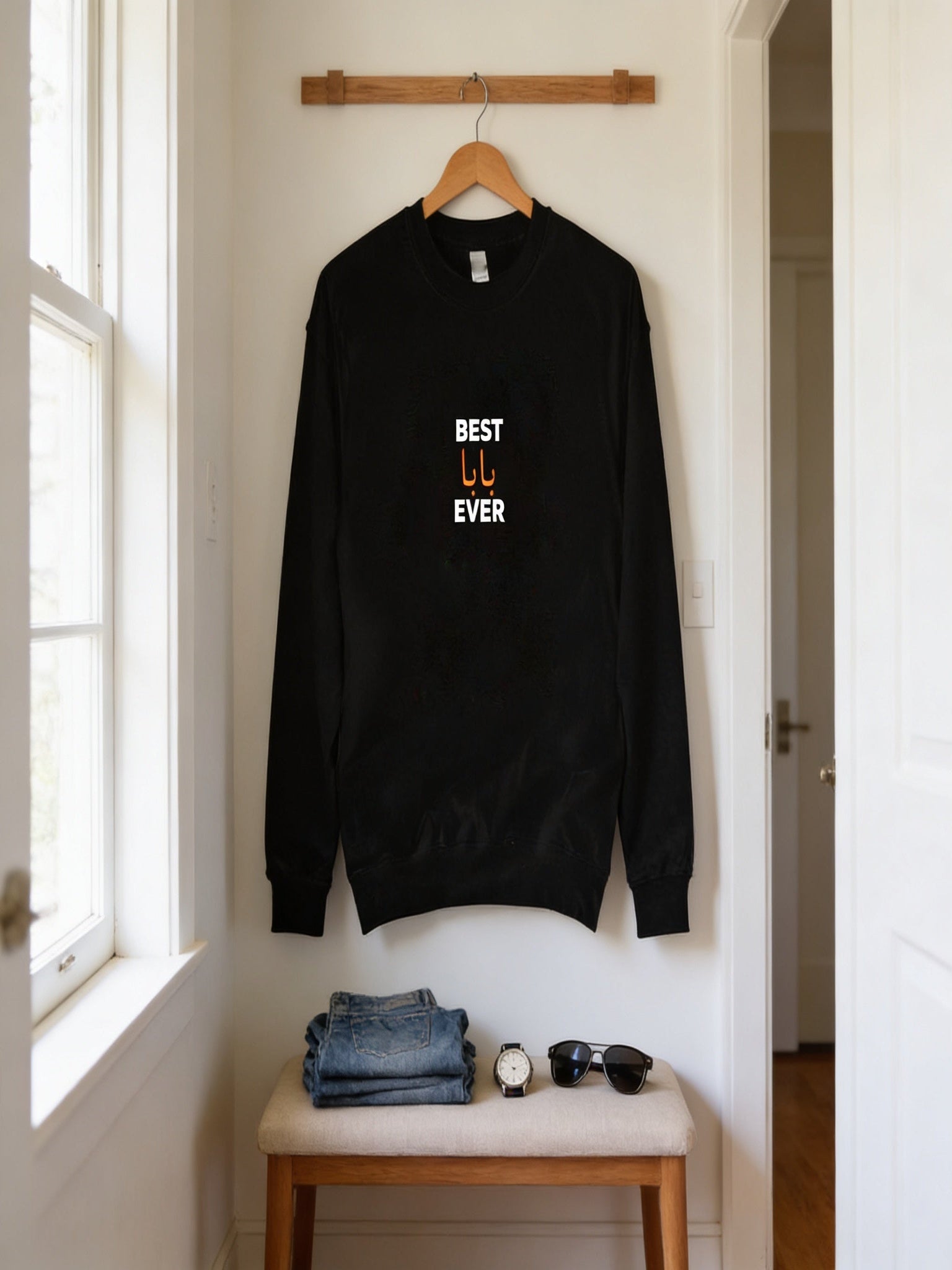 A black sweatshirt with the text "BEST U EVER" printed on it is hanging on a wooden hanger on a wall. Below the sweatshirt, there is a folded pair of jeans and a pair of sunglasses.