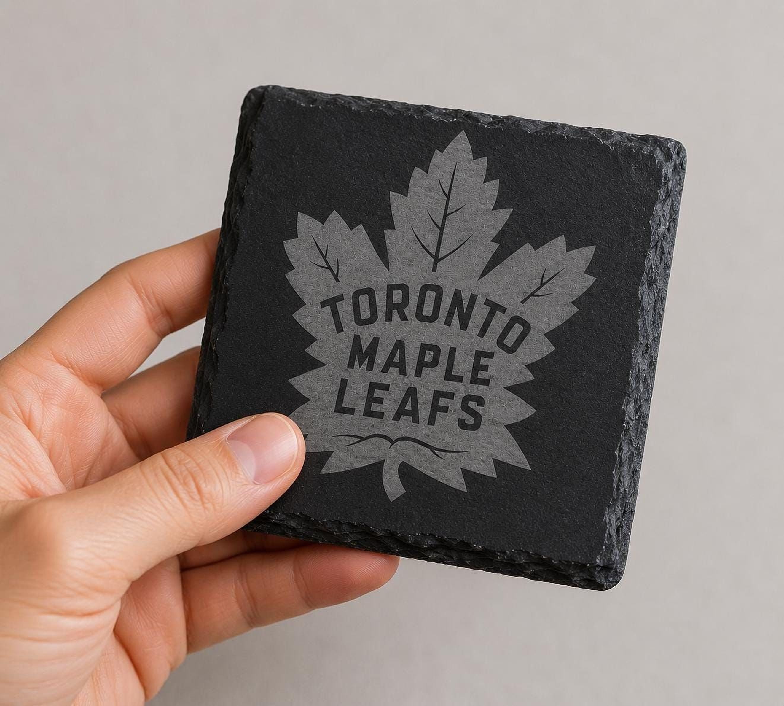A hand holding a black coaster with a maple leaf design and the text "TORONTO MAPLE LEAFS" printed on it.