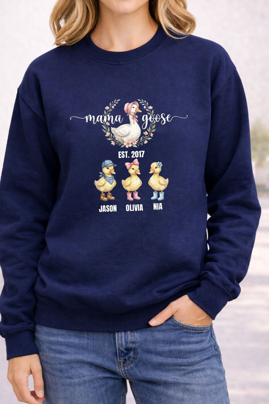 A woman wearing a navy blue sweatshirt with a graphic design featuring three cartoon ducks and the text "Mama Goose" above them.