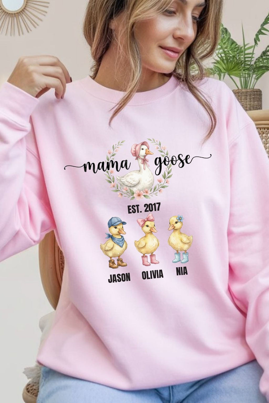 A woman wearing a pink sweatshirt with a graphic design featuring three cartoon ducks and the text "mama goose" and "Est. 2017 Jason Olivia Nia".