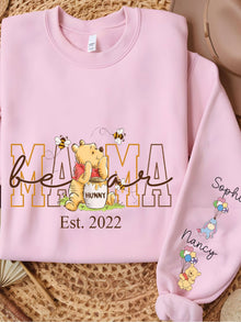 A pink sweatshirt with a Winnie the Pooh-themed design, featuring the character Winnie the Pooh and the text "Mama" and "Est. 2020". The sweatshirt also has a personalized embroidered sleeve with the names "Sophia" and "Nancy".