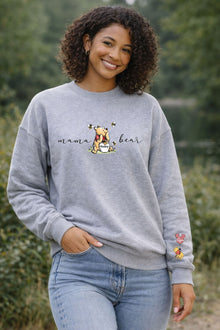 A smiling woman wearing a gray sweatshirt with a Winnie the Pooh graphic and the text "mama bear" printed on it, standing in a natural outdoor setting.