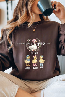 A woman wearing a brown sweatshirt with a graphic design featuring three cartoon ducks and the text "mama goose" and "Est. 2017 Jason Olivia Nia".