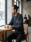 A man in a black sweatshirt is sitting at a table in a coffee shop, holding a cup of coffee and looking out the window.