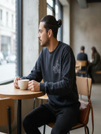 A man in a black sweatshirt is sitting at a table in a coffee shop, holding a cup of coffee and looking out the window.