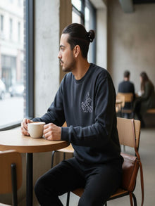 A man in a black sweatshirt is sitting at a table in a coffee shop, holding a cup of coffee and looking out the window.