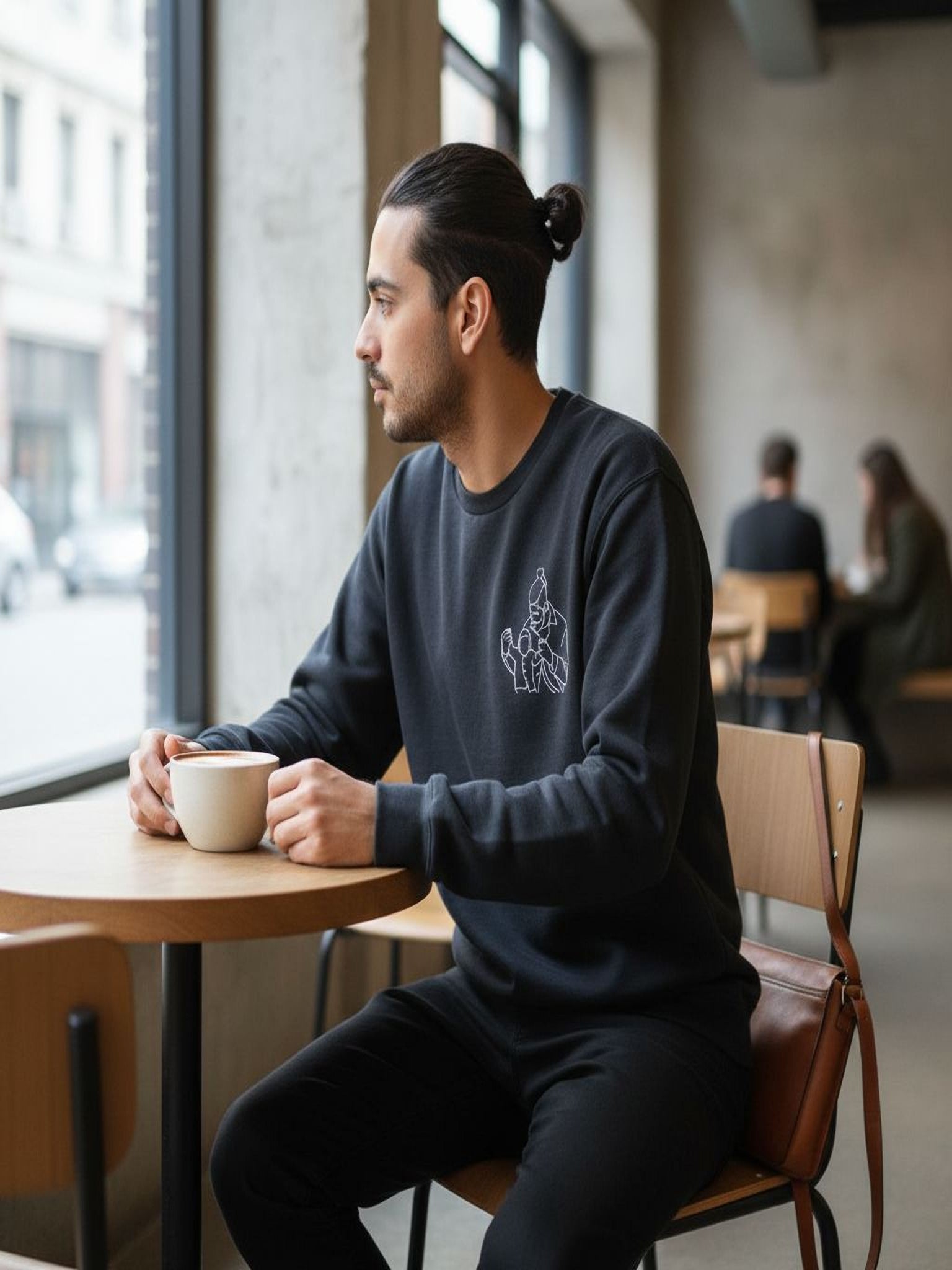 A man in a black sweatshirt is sitting at a table in a coffee shop, holding a cup of coffee and looking out the window.