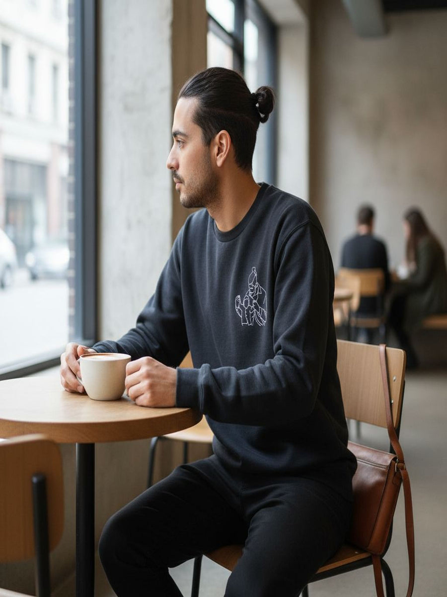 A man in a black sweatshirt is sitting at a table in a coffee shop, holding a cup of coffee and looking out the window.