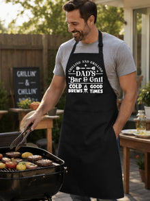 Dad’s Bar and Grill apron featuring funny BBQ typography design worn by man grilling outdoors in backyard.