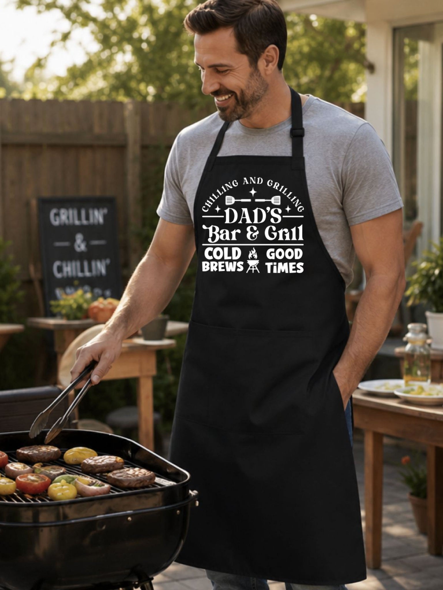 Dad’s Bar and Grill apron featuring funny BBQ typography design worn by man grilling outdoors in backyard.