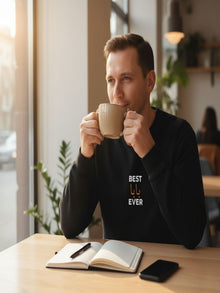 A man is sitting at a table in a cafe, holding a cup of coffee and looking out the window.