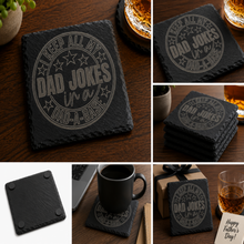 Black engraved slate coaster featuring the phrase "Keep All My Dad Jokes in a Dad-A-Base" displayed on a wooden tabletop beside a whiskey glass.