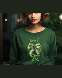 Dark green women's Christmas hoodie with festive Grinch Era graphic near tree