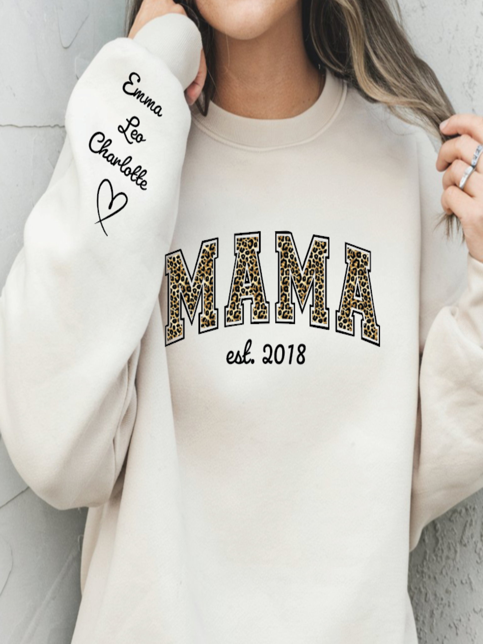 Person wearing a beige sweatshirt with 'MAMA est. 2018' text and white arm sleeve with 'Emma Leo Charlotte' text.
