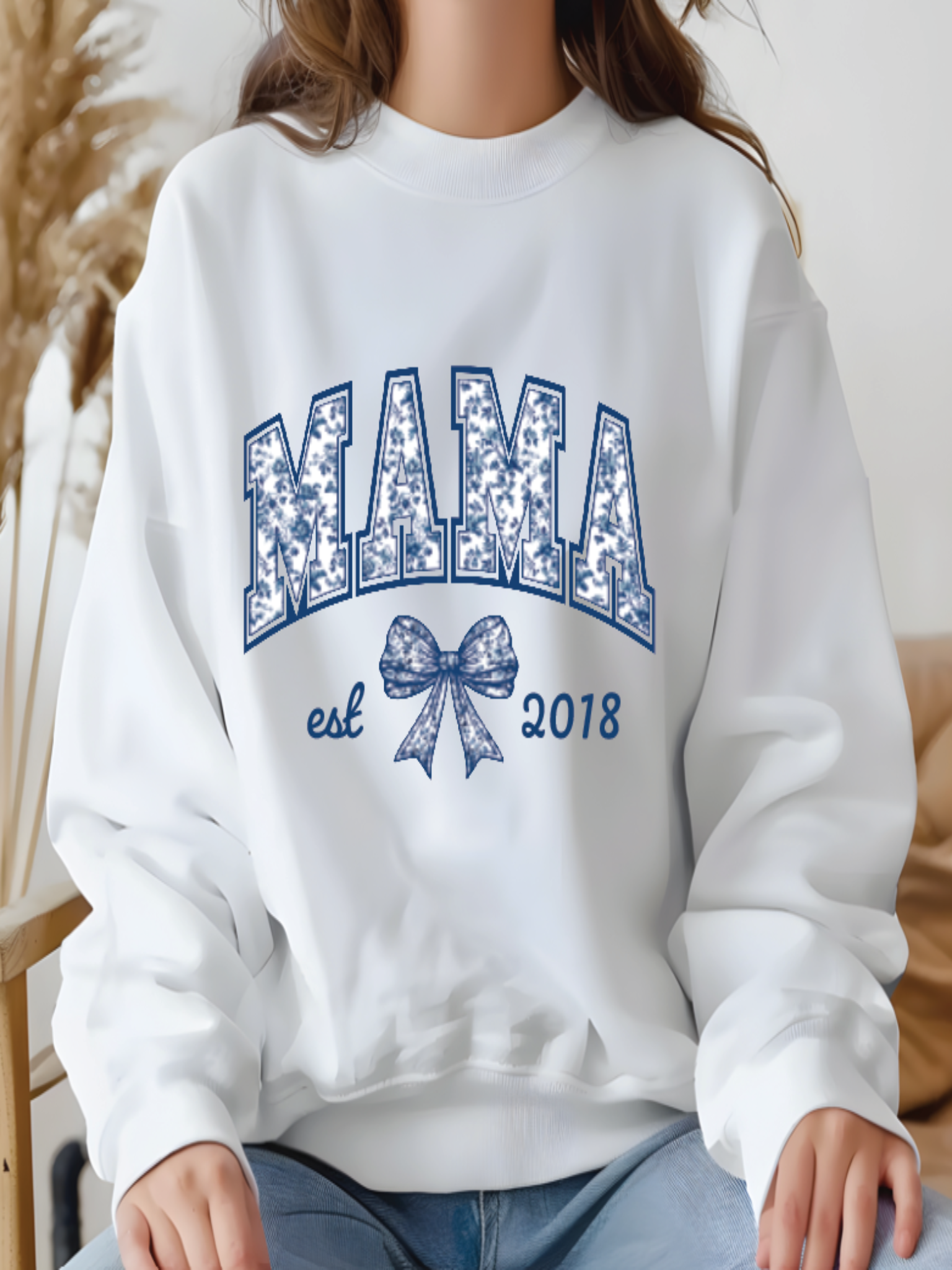 White sweatshirt with 'MAMA est 2018' text and bow design, worn by a person.