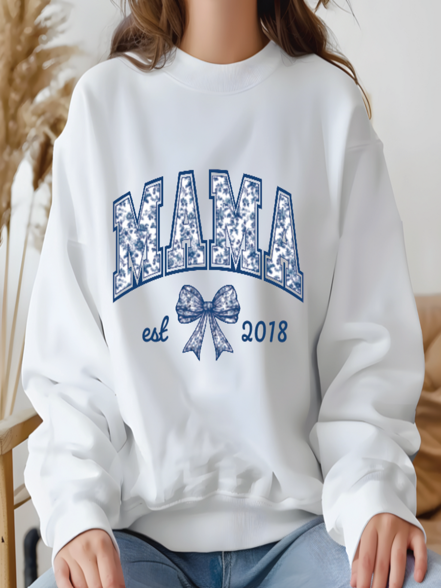 White sweatshirt with 'MAMA est 2018' text and bow design, worn by a person.
