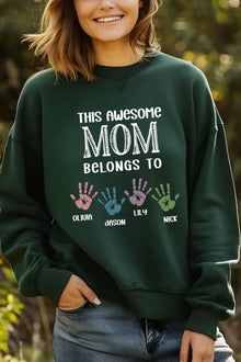 A woman wearing a green sweatshirt with text that says "This awesome MOM belongs to" and handprints of three different people.