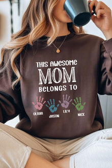A woman wearing a brown sweatshirt with a printed message that says "This awesome MOM belongs to" and handprints of three children's names below it.