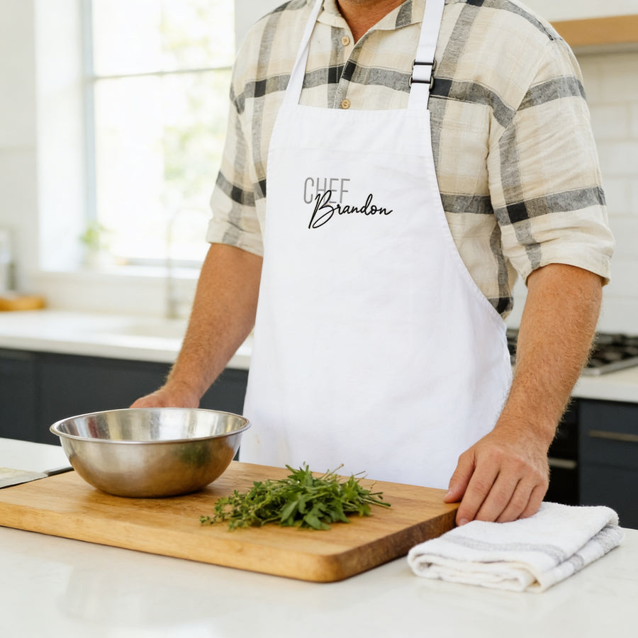 A man wearing a white apron with the name "Chef Brandon" is standing in a kitchen, preparing food on a wooden cutting board.