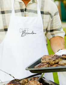A person wearing a white apron with the name "Chef Brandon" printed on it is holding a tray of grilled meat.