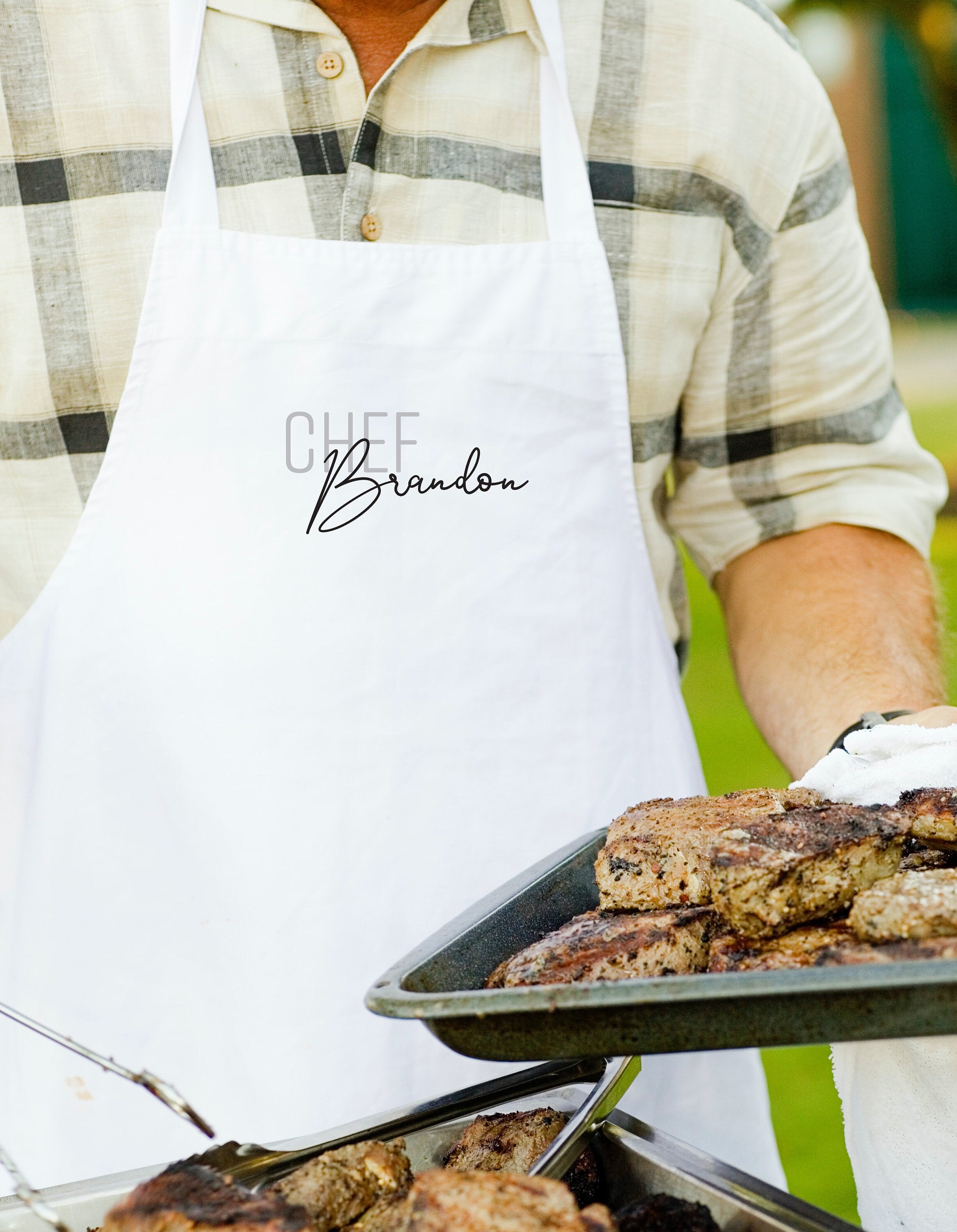 A person wearing a white apron with the name "Chef Brandon" printed on it is holding a tray of grilled meat.