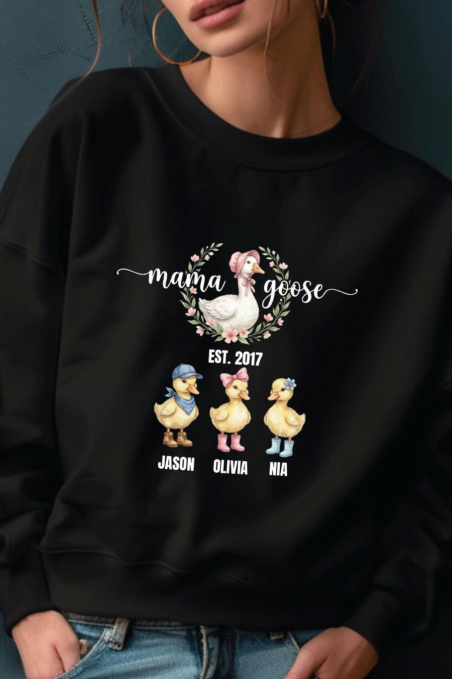 A woman wearing a black sweatshirt with a graphic design featuring three cartoon ducks and the text "mama goose" above them.