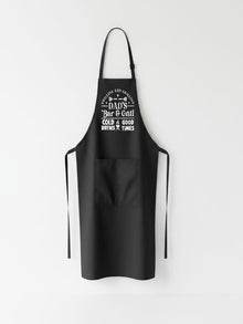 Dad’s Bar and Grill apron featuring funny BBQ typography design worn by man grilling outdoors in backyard.