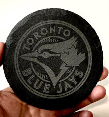A hand holding a black coaster with the logo of the Toronto Blue Jays baseball team printed on it.