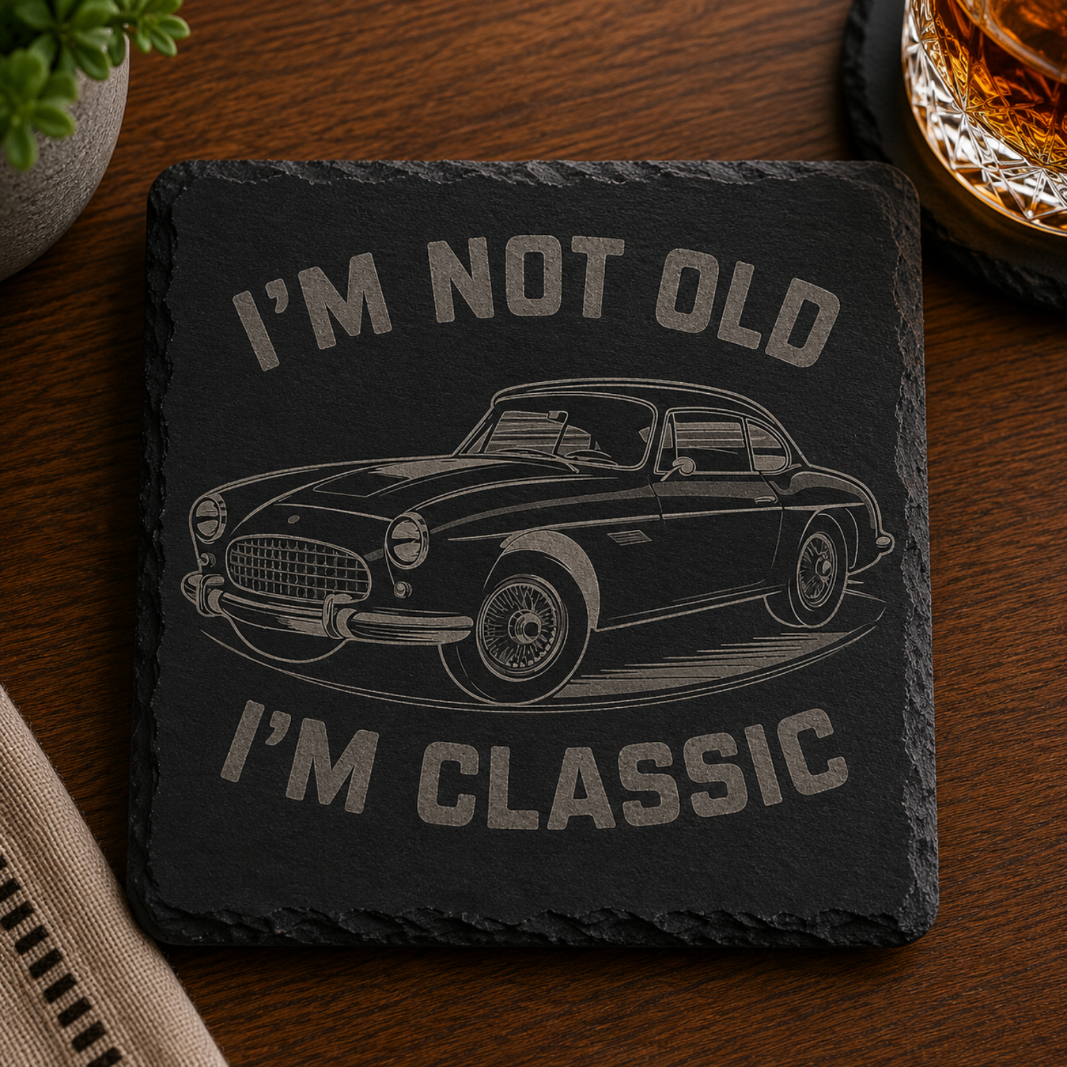 Engraved black slate coaster featuring a vintage classic car and the quote "I'm Not Old I'm Classic" displayed on a wooden tabletop.