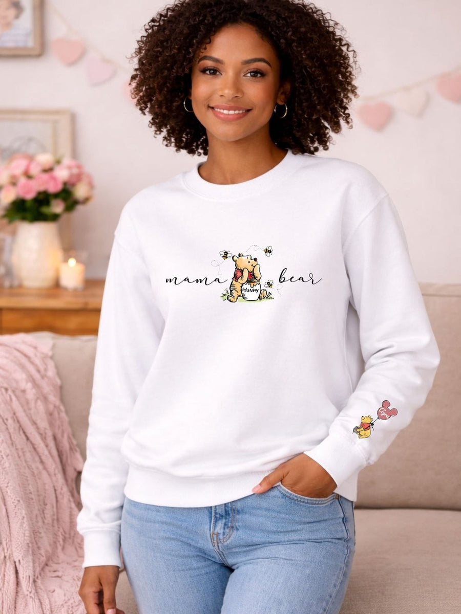 A woman with curly hair is smiling and wearing a white sweatshirt with a cartoon character and the text "mama bear" printed on it. She is standing in front of a pink blanket and a vase of flowers, with a couch visible in the background.