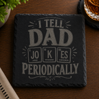 Engraved black slate coaster featuring the phrase "I Tell Dad Jokes Periodically" with a periodic table-inspired design, displayed on a wooden table beside a whiskey glass.