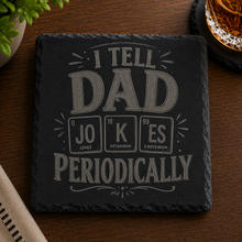 Engraved black slate coaster featuring the phrase "I Tell Dad Jokes Periodically" with a periodic table-inspired design, displayed on a wooden table beside a whiskey glass.