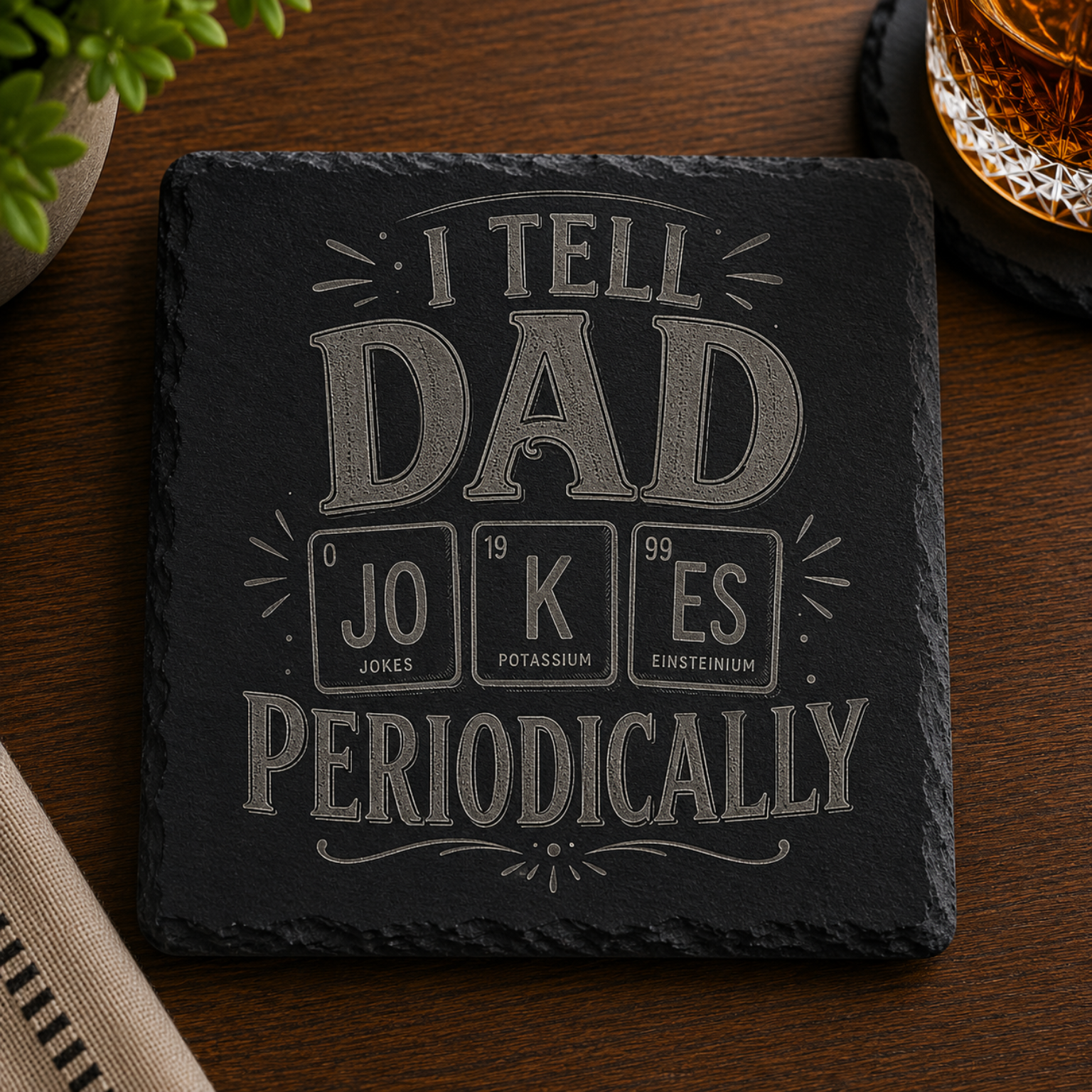 Engraved black slate coaster featuring the phrase "I Tell Dad Jokes Periodically" with a periodic table-inspired design, displayed on a wooden table beside a whiskey glass.