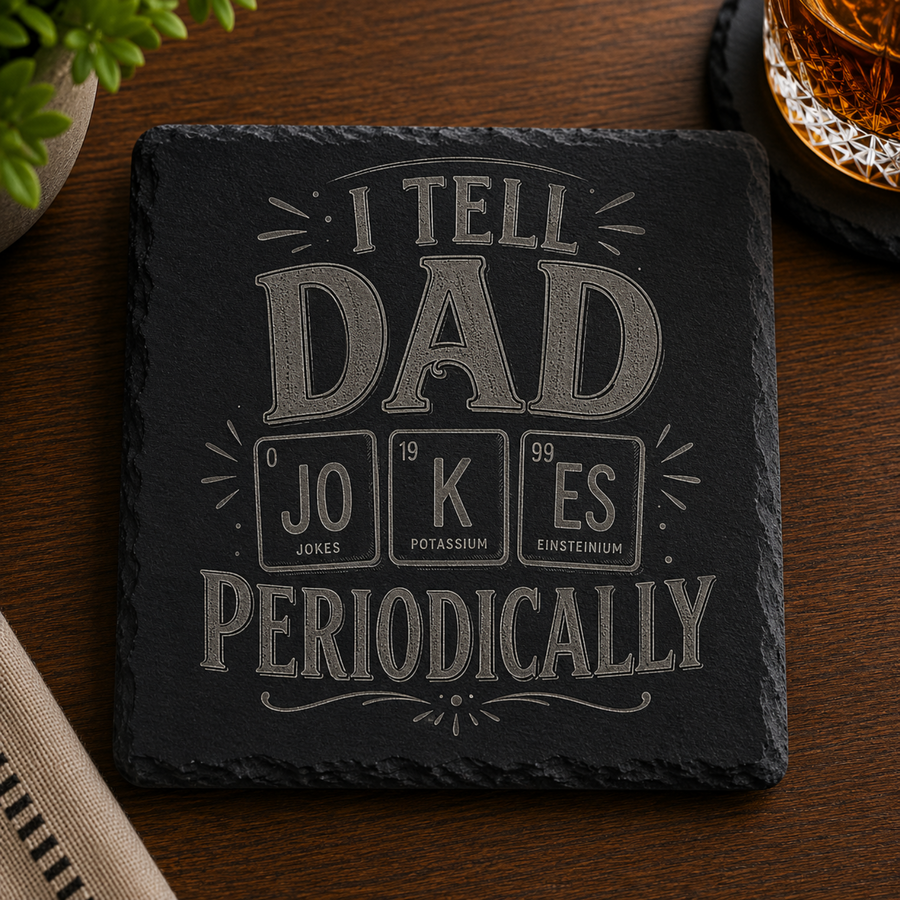 Engraved black slate coaster featuring the phrase "I Tell Dad Jokes Periodically" with a periodic table-inspired design, displayed on a wooden table beside a whiskey glass.
