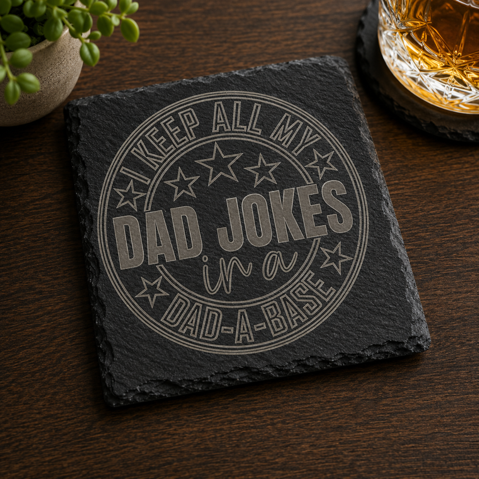 Black engraved slate coaster featuring the phrase "Keep All My Dad Jokes in a Dad-A-Base" displayed on a wooden tabletop beside a whiskey glass.