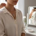 A woman wearing a necklace with a pendant stands in front of a mirror, with a white vase of flowers and a small bowl of jewelry visible in the background.