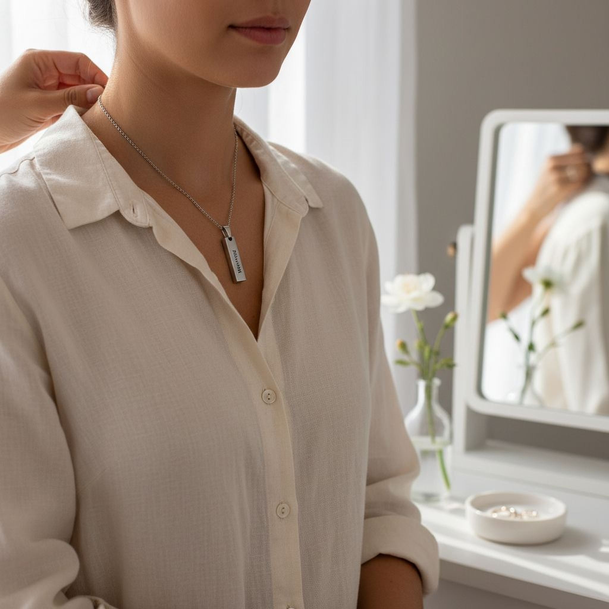 A woman wearing a necklace with a pendant stands in front of a mirror, with a white vase of flowers and a small bowl of jewelry visible in the background.