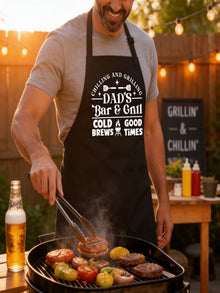 Dad’s Bar and Grill apron featuring funny BBQ typography design worn by man grilling outdoors in backyard.
